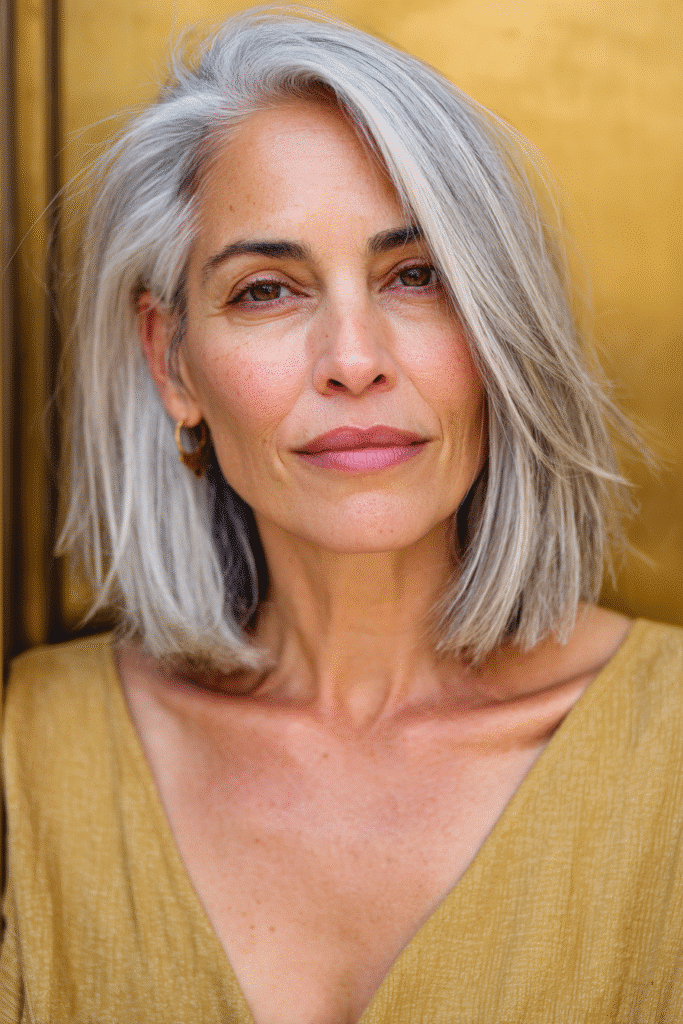 A vibrant American female model over 50 with a mature appearance, showcasing a youthful Classic Sleek Bob hairstyle with a straight, polished finish, captured in a close-up shot focusing on the hair, photographed against a seamless warm gold backdrop with a glossy sheen. She has subtle makeup with fine lines, an elegant outfit, realistic hair texture with silver highlights, warm skin tone, and a friendly expression, captured with soft natural lighting and an Instagram-style beauty vibe.