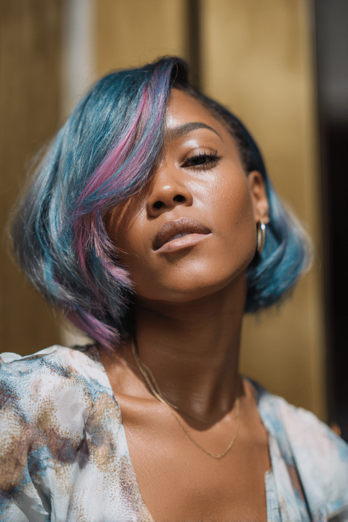 A vibrant female model in her 20s with a shoulder-length colored quick weave bob, styled with vibrant dye accents with blue and purple and also a pint layer, captured in a close-up shot focusing on the hair, photographed against a seamless warm gold backdrop with a glossy sheen. She has subtle makeup, a trendy outfit, a warm skin tone, and a confident expression, captured with warm natural lighting and a soft glow.