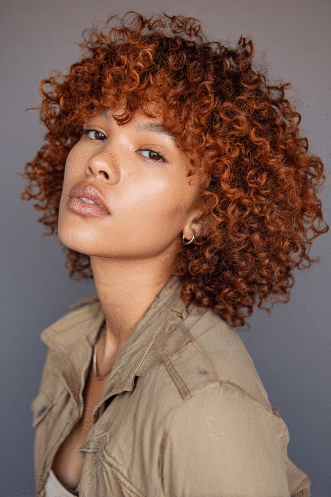 A vibrant young woman in her 20s with natural skin tone, showcasing a curly hairstyle for short hair with copper curls like Amandla Stenberg, featuring shiny copper-toned curls, captured in a close-up shot focusing on the hair, photographed against a seamless soft plum backdrop with a matte finish. She has subtle makeup, a trendy full-sleeve outfit, realistic hair texture, warm skin tone, and a confident expression, captured with soft natural lighting and an Instagram-style beauty vibe.