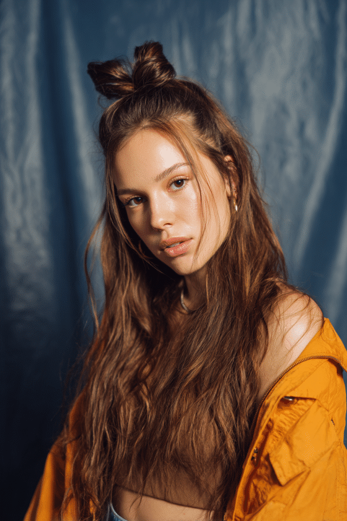 A vibrant female model in her 20s with shoulder-to-waist-length crown quick weave hair, styled with an elevated top section, captured in a close-up shot focusing on the hair, photographed against a seamless soft navy backdrop with a glossy sheen. She has subtle makeup, a trendy outfit, a warm skin tone, and a confident expression, captured with warm natural lighting and a soft glow.