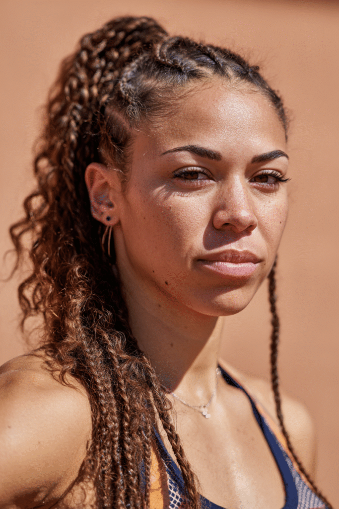 A vibrant female athlete in her 20s with very long hair extending past the waist, styled in curved braids and a pony with a secure tail, captured in a close-up shot focusing on the hair, photographed against a seamless warm peach backdrop with a matte finish. She has subtle makeup, a volleyball uniform, a warm skin tone, and a confident expression, captured with bright natural lighting and a sporty vibe.