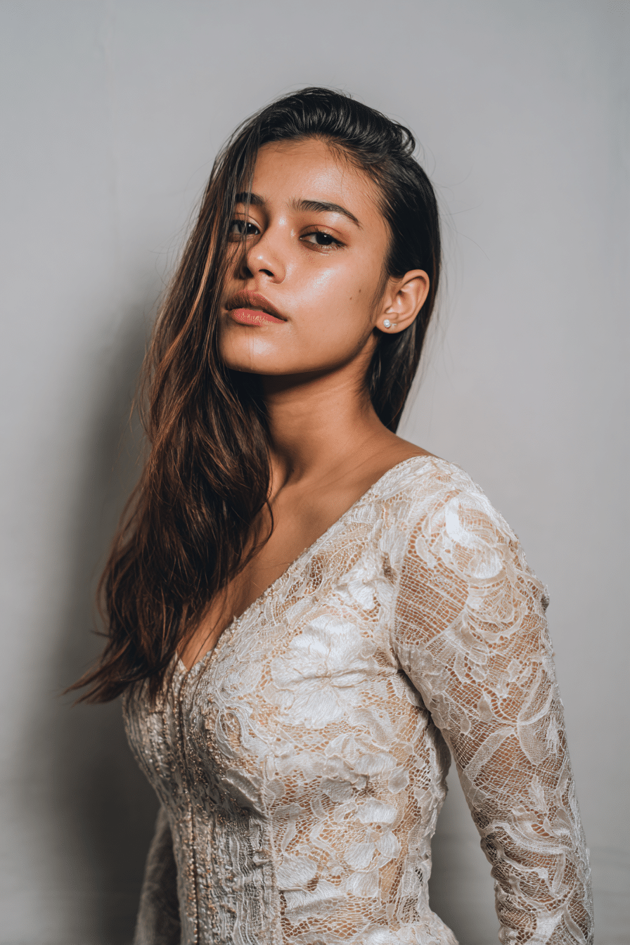 A gorgeous young woman with a natural look displaying sleek slicked-back hair with a deep side part in a light golden shade, wearing an elegant full-length dress with lace long sleeves, radiant expression, warm skin tone, against a seamless soft linen backdrop with a matte finish.