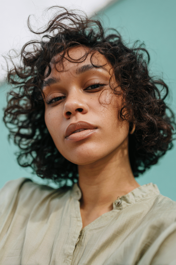 A vibrant young woman in her 20s with natural skin tone, showcasing a curly hairstyle for short hair with defined bob curls like Ruth Negga, featuring tight, structured curls, captured in a close-up shot focusing on the hair, photographed against a seamless light teal backdrop with a glossy sheen. She has subtle makeup, a trendy full-sleeve outfit, realistic hair texture, warm skin tone, and a confident expression, captured with soft natural lighting and an Instagram-style beauty vibe.