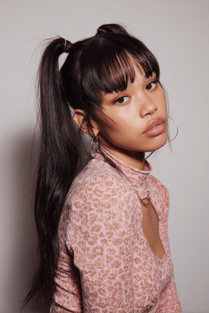 A young American female model with a warm skin tone and friendly expression, showcasing a Double Ponytail with Blunt Bob Bangs hairstyle. The hair features natural clip-in bangs on a straight ponytail, set against a soft gray fabric backdrop with minimal texture. She wears subtle makeup and a trendy casual outfit, captured in soft natural lighting for an Instagram-style beauty photo.
