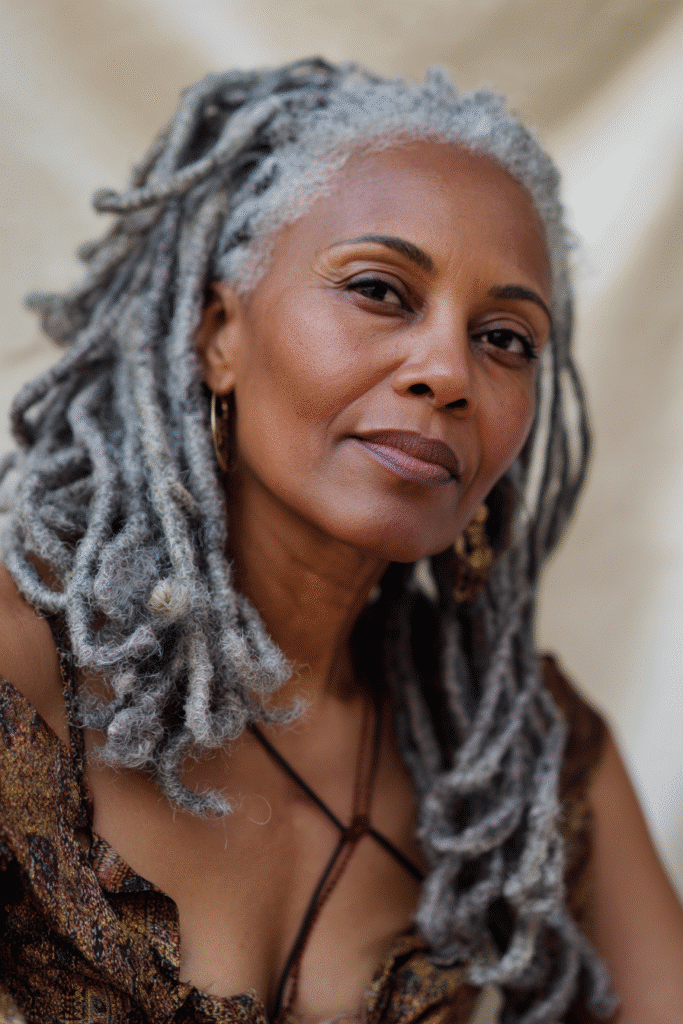 An elegant Black woman over 50 with a warm skin tone and friendly expression, showcasing elegant gray dreadlocks braids hairstyle for older Black women over 50. The hair is gray, set against a seamless white backdrop with minimal texture. She wears subtle makeup and an elegant outfit suitable for mature women, captured in soft natural lighting for an Instagram-style beauty photo.