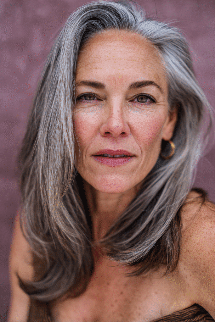 A vibrant American female model over 50 with a mature appearance, showcasing a youthful Face-Framing Long Layers hairstyle with soft framing strands, captured in a close-up shot focusing on the hair, photographed against a seamless warm violet backdrop with a matte finish. She has subtle makeup with visible aging, an elegant outfit, realistic hair texture with silver accents, warm skin tone, and a friendly expression, captured with soft natural lighting and an Instagram-style beauty vibe.