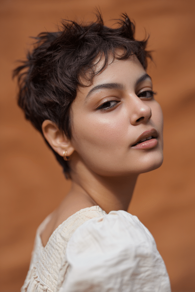 A gorgeous young female model in her 20s with natural skin tone, showcasing a prom hairstyle for short hair with a faux hawk pixie and volume, featuring added height at the top for edge, captured in a close-up shot focusing on the hair, photographed against a seamless warm cinnamon backdrop with silky texture. She has subtle makeup, an elegant full-shirt prom dress, realistic hair texture, warm skin tone, and a friendly expression, captured with soft natural lighting and an Instagram-style beauty vibe.