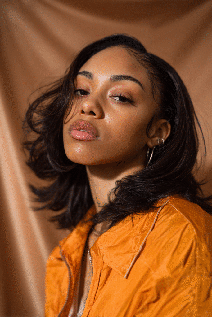 A vibrant female model in her 20s with shoulder-to-waist-length feathered quick weave hair, styled with light layered ends, captured in a close-up shot focusing on the hair, photographed against a seamless warm bronze backdrop with a satin finish. She has subtle makeup, a trendy outfit, a warm skin tone, and a confident expression, captured with warm natural lighting and a soft glow.