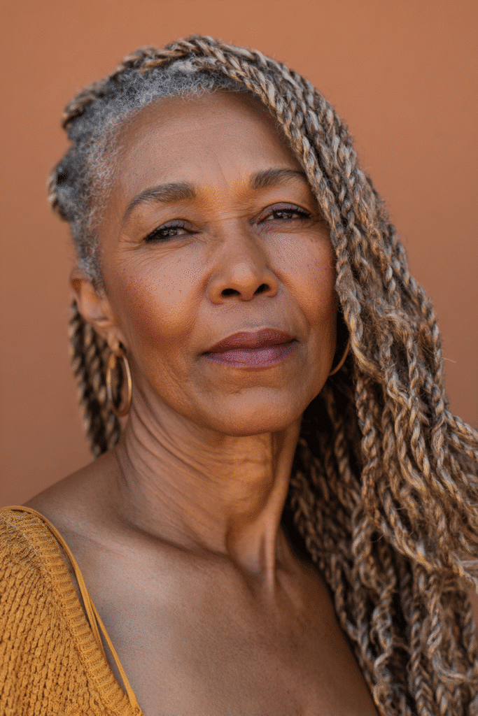 An elegant Black woman over 50 with a warm skin tone and friendly expression, showcasing graceful side-swept braids hairstyle for older Black women over 50. The hair is natural black, set against a natural forest scene backdrop with minimal texture. She wears subtle makeup and an elegant outfit suitable for mature women, captured in soft natural lighting for an Instagram-style beauty photo.