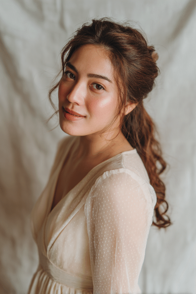 A gorgeous young bride with a natural look displaying elegant half-up twists with flowing long hair in a light caramel shade, wearing an elegant full-length wedding dress with chiffon long sleeves, radiant expression, warm skin tone, against a seamless soft linen backdrop with a matte finish.
