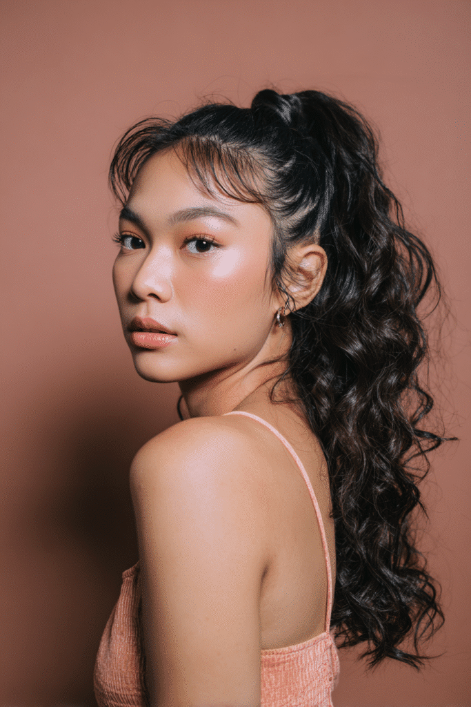 A stunning young woman in her 20s with natural skin tone, showcasing a homecoming hairstyle for medium hair with a high ponytail with curls, featuring a high ponytail with cascading curls using medium-length hair, captured in a close-up shot focusing on the hair, photographed against a seamless warm peach backdrop with a matte finish. She has subtle makeup, a chic homecoming dress, realistic hair texture, warm skin tone, and a confident expression, captured with soft natural lighting and an Instagram-style beauty vibe.