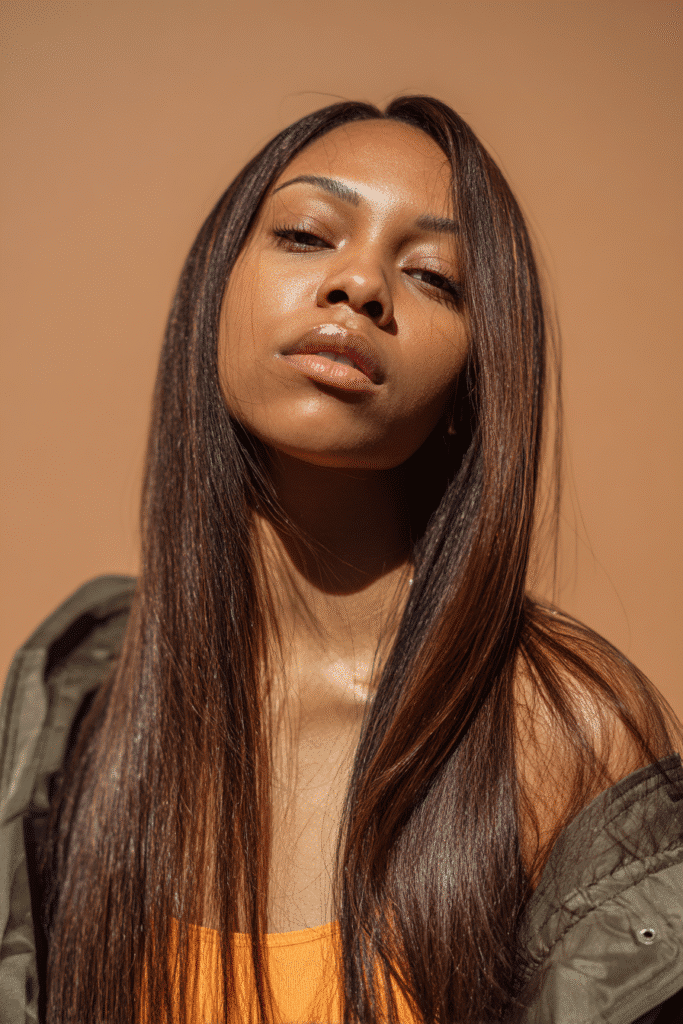 A vibrant female model in her 20s with long straight quick weave hair extending past the shoulders, styled with a smooth finish, captured in a close-up shot focusing on the hair, photographed against a seamless warm amber backdrop with a matte finish. She has subtle makeup, a trendy outfit, a warm skin tone, and a confident expression, captured with warm natural lighting and a soft glow.