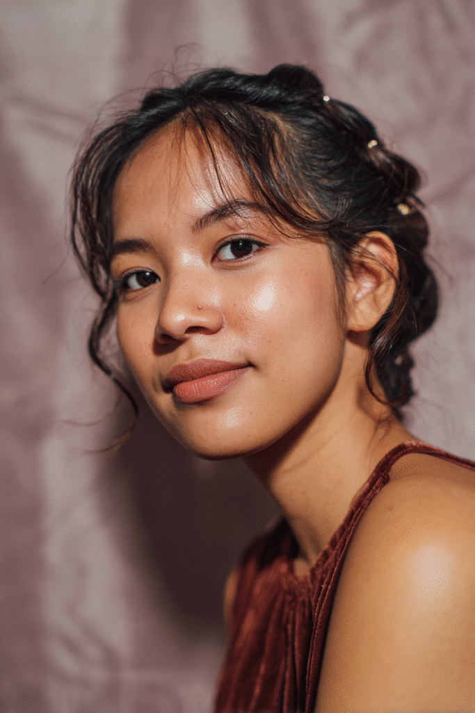 A gorgeous young female model in her 20s with natural skin tone, showcasing a prom hairstyle for short hair with a low twisted updo and hair pins, featuring a sophisticated pinned design, captured in a close-up shot focusing on the hair, photographed against a seamless soft mauve backdrop with glossy sheen. She has subtle makeup, an elegant full-shirt prom dress, realistic hair texture, warm skin tone, and a friendly expression, captured with soft natural lighting and an Instagram-style beauty vibe.