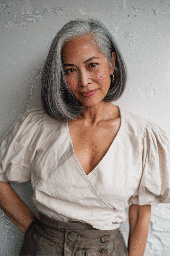 An elegant woman over 50 with a warm skin tone and serene expression, showcasing Lucy Liu's sleek bob hairstyle for bob hairstyles for women over 50. The hair is a straight sleek bob with gloss, colored jet black, styled with a satin kimono top. Captured in soft natural lighting with subtle makeup, set against a seamless white backdrop with minimal texture for an Instagram-style beauty photo.