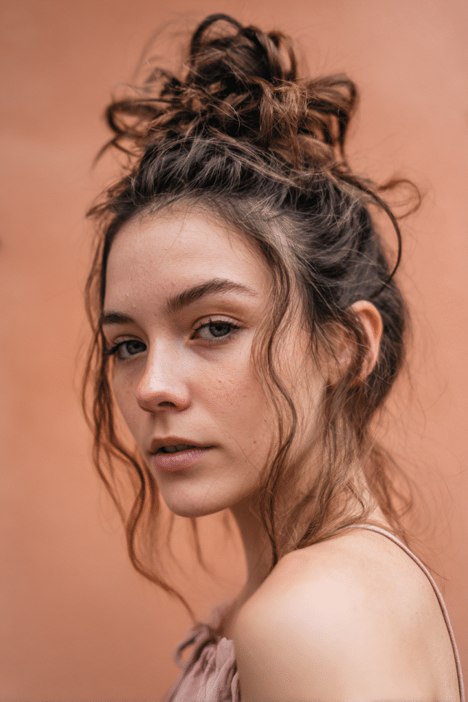 A stunning young woman in her 20s with natural skin tone, showcasing a homecoming hairstyle with a messy bun, featuring a high bun with loose strands, captured in a close-up shot focusing on the hair, photographed against a seamless warm coral backdrop with a smooth texture. She has subtle makeup, a chic homecoming dress, realistic hair texture, warm skin tone, and a confident expression, captured with soft natural lighting and an Instagram-style beauty vibe.