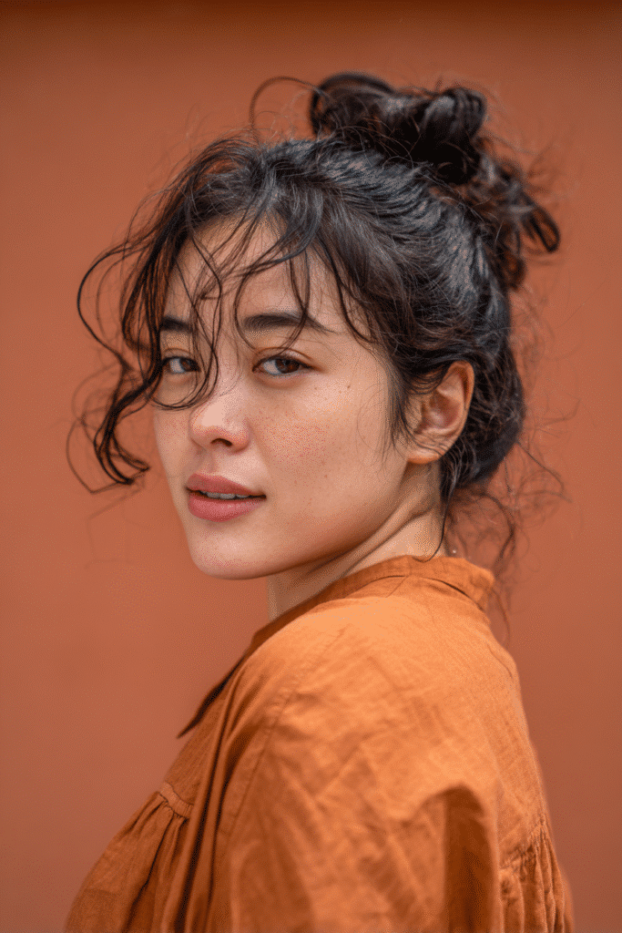 A vibrant young female model in her 20s with natural skin tone, showcasing a 90s messy updo with a messy bun with clip-in extensions, featuring added volume from extensions, captured in a close-up shot focusing on the hair, photographed against a seamless warm terracotta backdrop with a satin finish. She has subtle makeup, a retro full-shirt 90s outfit, realistic hair texture, warm skin tone, and a friendly expression, captured with soft natural lighting and an Instagram-style beauty vibe.