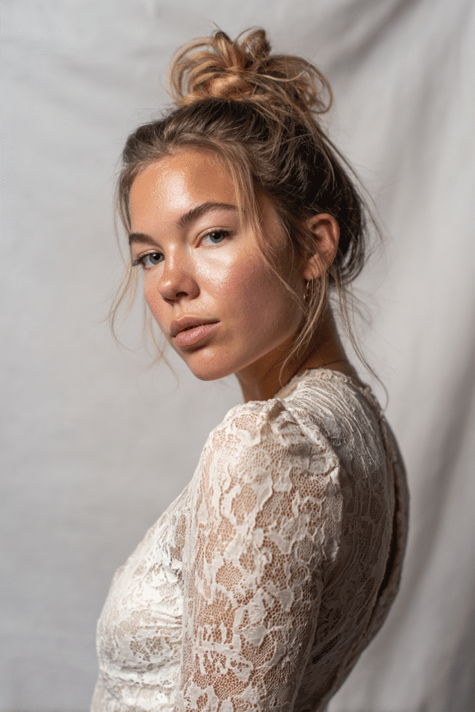 A gorgeous young woman with a natural look featuring a textured messy bun in a light caramel shade, wearing an elegant full-length dress with lace long sleeves, radiant expression, warm skin tone, against a seamless soft linen backdrop with a matte finish.