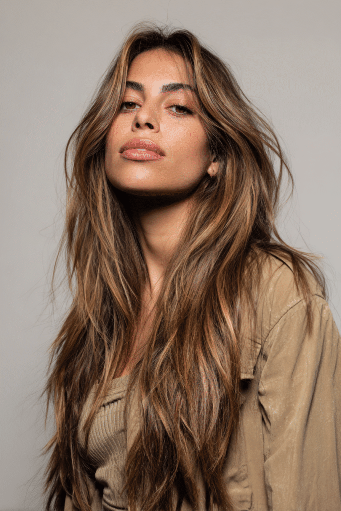 A vibrant female model in her 20s with shoulder-to-waist-length hair, styled in a multi-dimensional brunette balayage with layered highlights, captured in a close-up shot focusing on the hair, photographed against a seamless light gray backdrop with a smooth surface. She has subtle makeup, a trendy outfit, a warm skin tone, and a confident expression, captured with warm natural lighting and a soft glow.