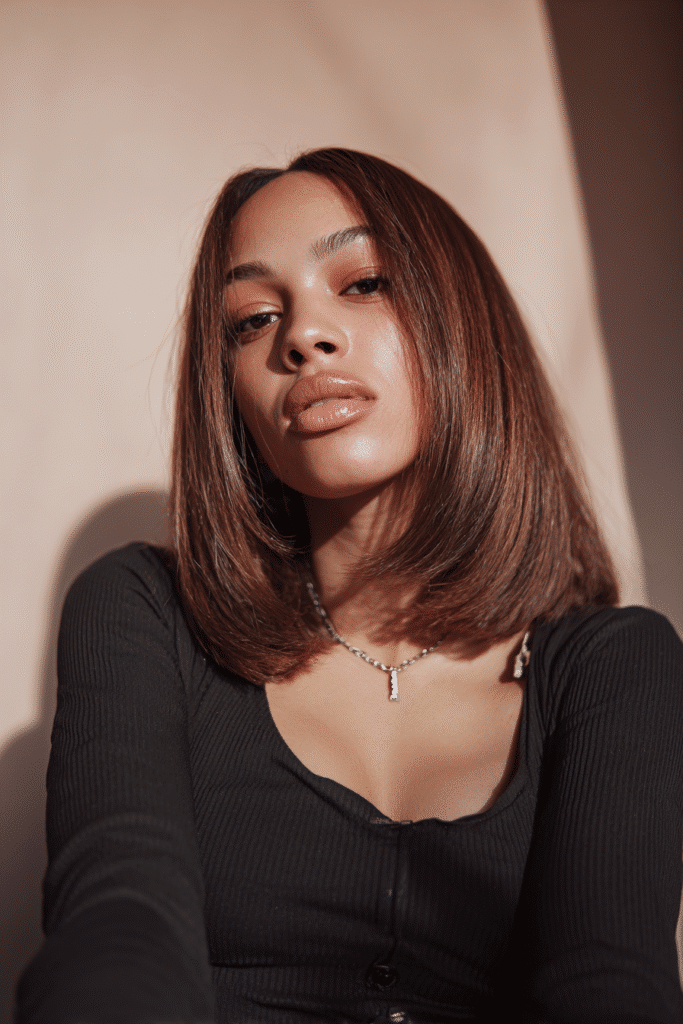 A vibrant female model in her 20s with shoulder-to-waist-length ombré quick weave hair, styled with a gradient color transition, captured in a close-up shot focusing on the hair, photographed against a seamless light coral backdrop with a smooth surface. She has subtle makeup, a trendy outfit, a warm skin tone, and a confident expression, captured with warm natural lighting and a soft glow.
