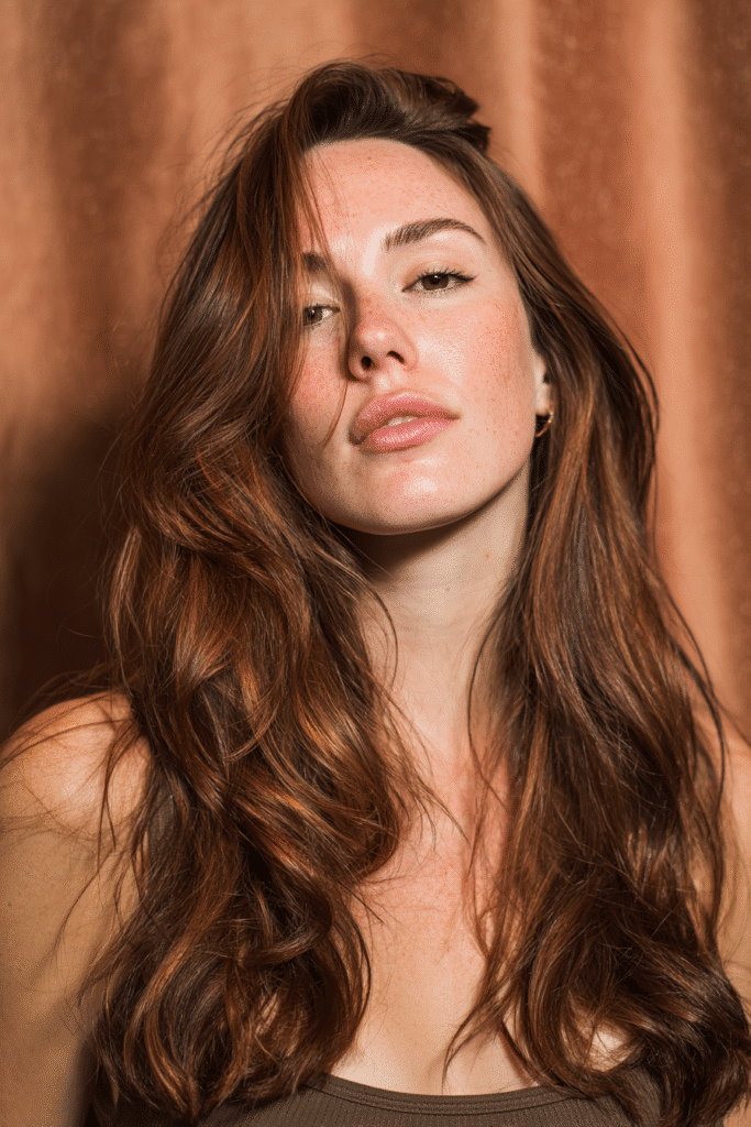 A vibrant female model in her 20s with shoulder-to-waist-length hair, styled in a rose brown balayage with pinkish-brown highlights, captured in a close-up shot focusing on the hair, photographed against a seamless warm bronze backdrop with a satin finish. She has subtle makeup, a trendy outfit, a warm skin tone, and a confident expression, captured with warm natural lighting and a soft glow.