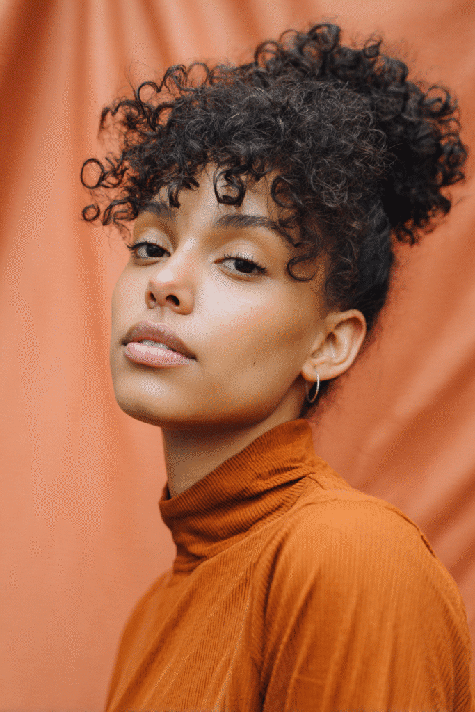 A vibrant young woman in her 20s with natural skin tone, showcasing a curly hairstyle for short hair with a short afro like Jodie Turner-Smith, featuring voluminous natural texture, captured in a close-up shot focusing on the hair, photographed against a seamless warm coral backdrop with a smooth texture. She has subtle makeup, a trendy full-sleeve outfit, realistic hair texture, warm skin tone, and a confident expression, captured with soft natural lighting and an Instagram-style beauty vibe.