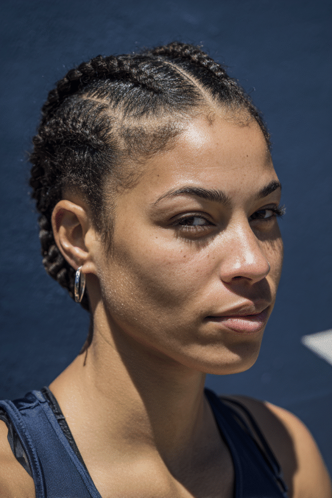 A vibrant female athlete in her 20s with short hair, styled in short hair cornrows with a tight, protective weave, captured in a close-up shot focusing on the hair, photographed against a seamless soft navy backdrop with a glossy sheen. She has subtle makeup, a volleyball uniform, a warm skin tone, and a confident expression, captured with bright natural lighting and a sporty vibe.