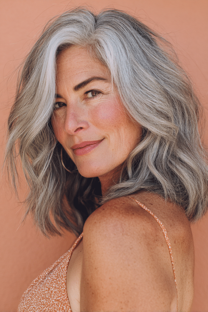 A vibrant American female model over 50 with a mature appearance, showcasing a youthful Shoulder-Length Waves hairstyle with loose, beachy curls for a fresh vibe, captured in a close-up shot focusing on the hair, photographed against a seamless light coral backdrop with a smooth surface. She has subtle makeup with fine lines, an elegant outfit, realistic hair texture with silver accents, warm skin tone, and a friendly expression, captured with soft natural lighting and an Instagram-style beauty vibe.
