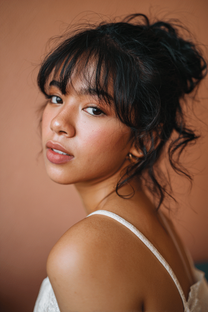 A stunning young female model in her 20s with natural skin tone, showcasing a messy bun hairstyle for wedding with a side-swept messy bun with bangs, featuring soft bangs and a side-swept style, captured in a close-up shot focusing on the hair, photographed against a seamless warm rose backdrop with a silky texture. She has subtle wedding makeup, an elegant full-sleeve wedding gown, realistic hair texture, warm skin tone, and a serene expression, captured with soft natural lighting and an Instagram-style beauty vibe.