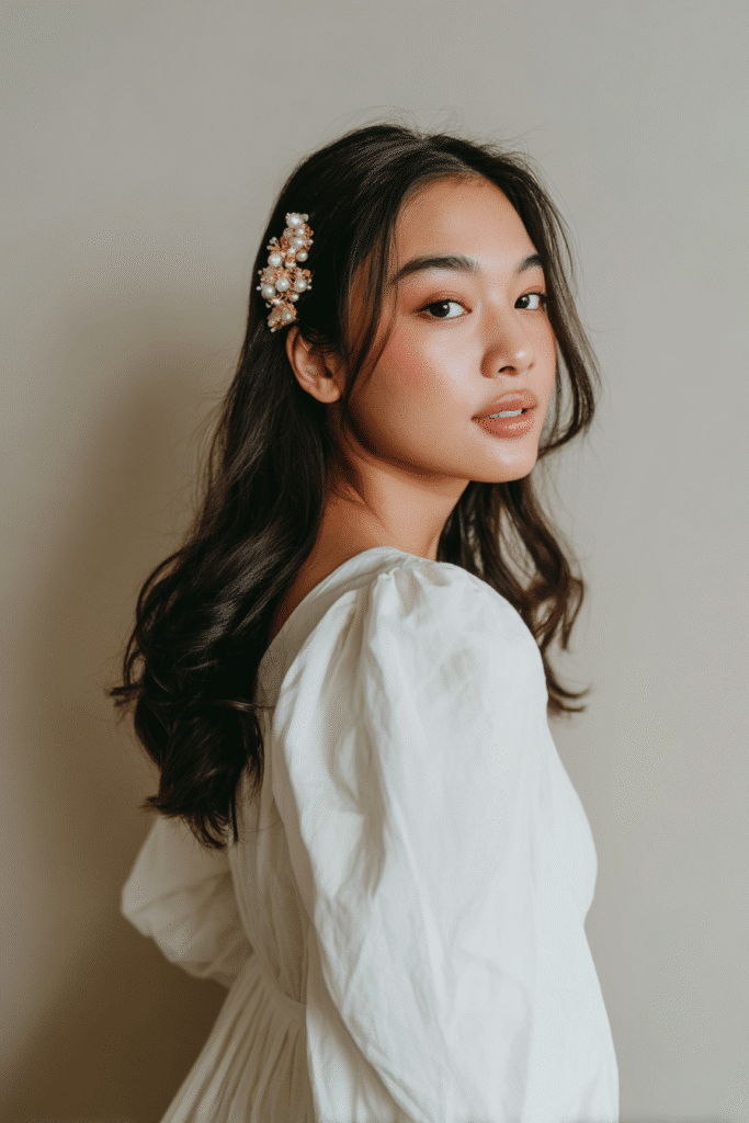 A gorgeous young bride with a natural look featuring soft side-swept waves with a decorative pearl clip in a light golden shade, wearing an elegant full-length wedding dress with chiffon long sleeves, radiant expression, warm skin tone, against a seamless soft linen backdrop with a matte finish.