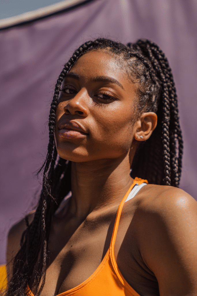 A vibrant female athlete in her 20s with shoulder-to-waist-length hair, styled in sleek Ghana braids with a tight, protective pattern, captured in a close-up shot focusing on the hair, photographed against a seamless warm violet backdrop with a matte finish. She has subtle makeup, a volleyball uniform, a warm skin tone, and a confident expression, captured with bright natural lighting and a sporty vibe.
