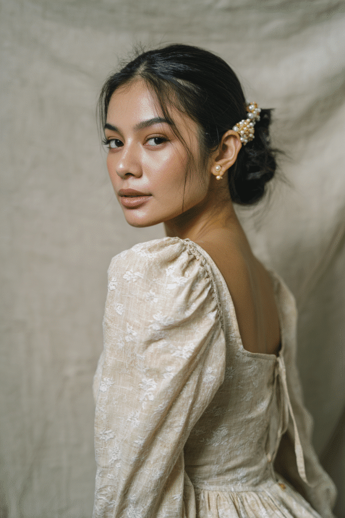A gorgeous young woman with a natural look featuring a sleek bun with pearl hair accessories in a light golden shade, wearing an elegant full-length dress with lace long sleeves, radiant expression, warm skin tone, against a seamless soft linen backdrop with a matte finish.