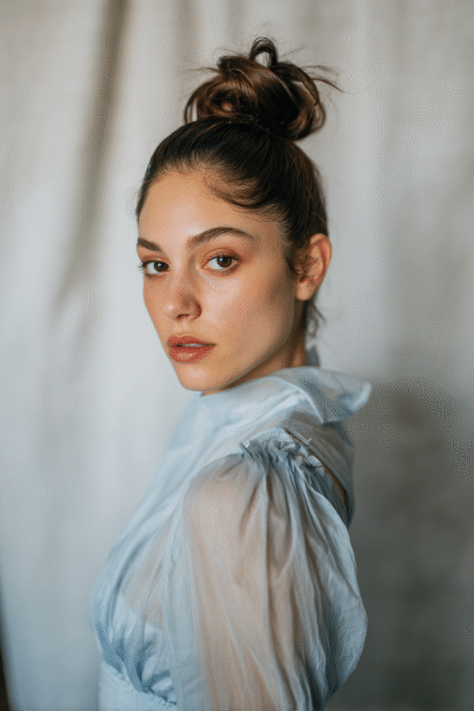 A gorgeous young woman with a natural look featuring a sleek bun with a voluminous crown in a soft hazelnut shade, wearing an elegant full-length dress with chiffon long sleeves, radiant expression, warm skin tone, against a seamless soft linen backdrop with a matte finish.