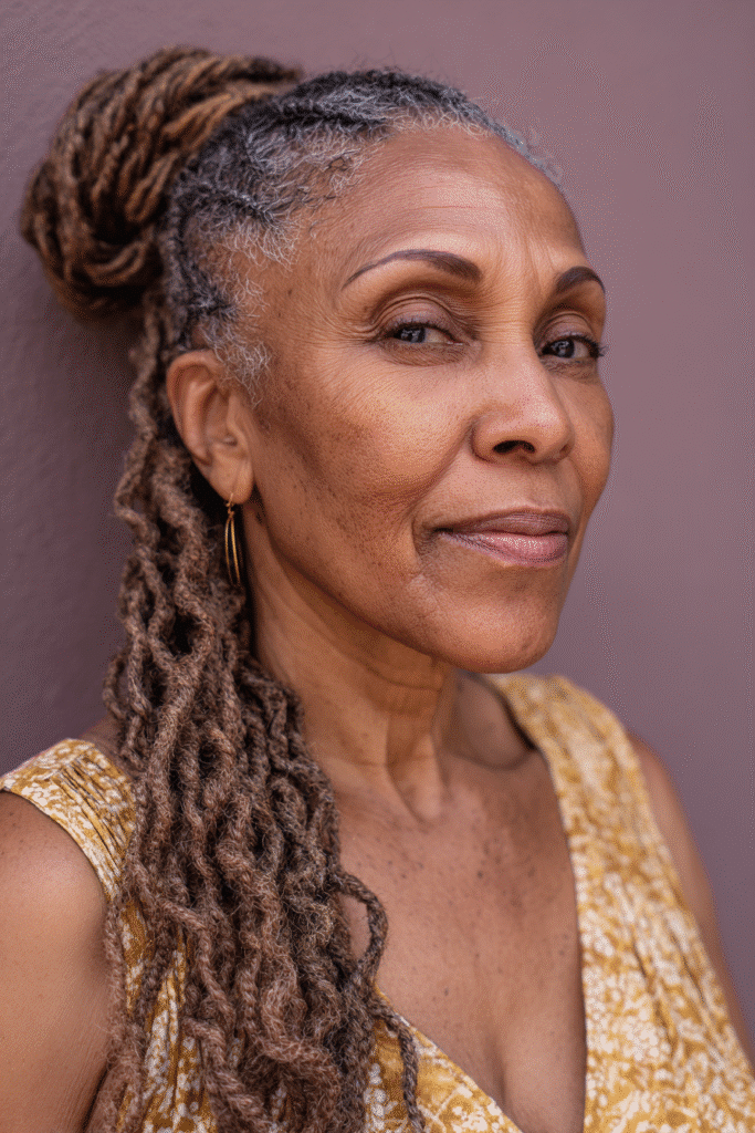 An elegant Black woman over 50 with a warm skin tone and friendly expression, showcasing twist together braided ponytail hairstyle for older Black women over 50. The hair is natural black, set against a soft gray fabric backdrop with minimal texture. She wears subtle makeup and an elegant outfit suitable for mature women, captured in soft natural lighting for an Instagram-style beauty photo.
