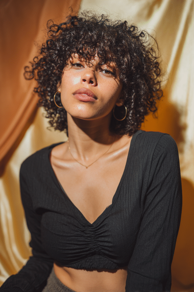 A vibrant young woman in her 20s with natural skin tone, showcasing a curly hairstyle for short hair with voluminous coils like Viola Davis, featuring full, textured coils, captured in a close-up shot focusing on the hair, photographed against a seamless warm amber backdrop with a silky texture. She has subtle makeup, a trendy full-sleeve outfit, realistic hair texture, warm skin tone, and a confident expression, captured with soft natural lighting and an Instagram-style beauty vibe.