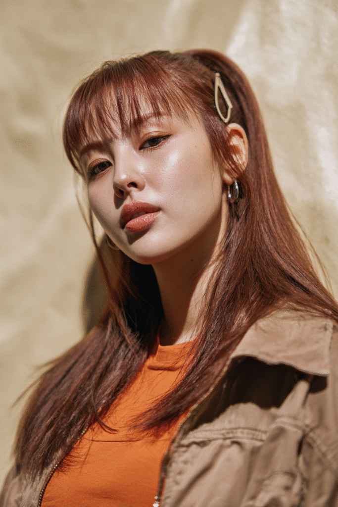 A young American female model with a warm skin tone and friendly expression, showcasing a Voluminous Ponytail with Layered Fringe hairstyle. The hair features side-swept bangs on a straight ponytail, set against a seamless white backdrop with minimal texture. She wears subtle makeup and a trendy casual outfit, captured in soft natural lighting for an Instagram-style beauty photo.