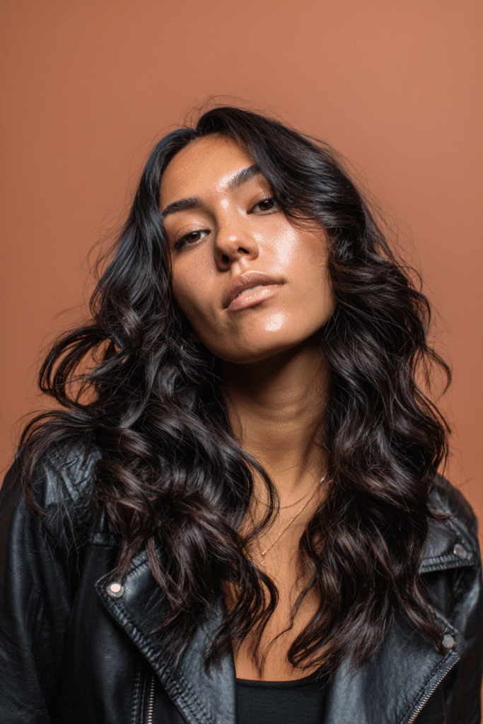 A vibrant female model in her 20s with shoulder-to-waist-length wavy quick weave hair, styled with natural loose waves, captured in a close-up shot focusing on the hair, photographed against a seamless warm plum backdrop with a satin finish. She has subtle makeup, a trendy outfit, a warm skin tone, and a confident expression, captured with warm natural lighting and a soft glow.