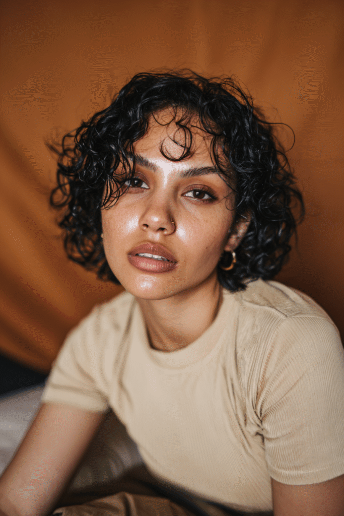 A vibrant young woman in her 20s with natural skin tone, showcasing a curly hairstyle for short hair with a wet look bob like Jourdan Dunn, featuring a sleek, wet-effect bob, captured in a close-up shot focusing on the hair, photographed against a seamless warm bronze backdrop with a satin finish. She has subtle makeup, a trendy full-sleeve outfit, realistic hair texture, warm skin tone, and a confident expression, captured with soft natural lighting and an Instagram-style beauty vibe.