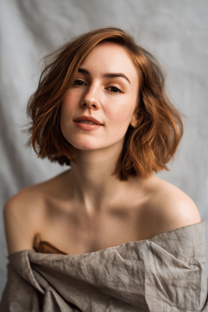 Young woman with shoulder-length angled bob, textured bangs, caramel brown hair, soft gray backdrop.