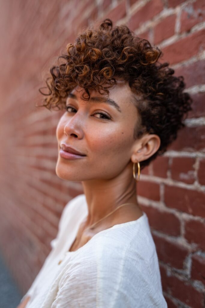 Woman with short 3c asymmetrical curly bob, chestnut brown hair, urban brick backdrop.
