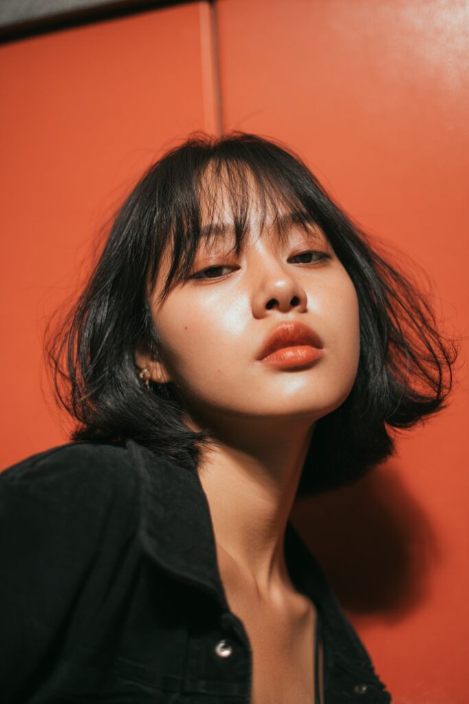 A stunning young woman in her 20s with natural skin tone, showcasing a bob hairstyle for going out featuring a blunt bob with flipped ends, featuring a blunt bob with flipped ends, captured in a close-up shot focusing on the hair, photographed against a seamless rich burgundy backdrop with a satin texture for optimal color contrast. She has subtle makeup, a chic evening outfit, realistic hair texture, warm skin tone, and a confident expression, captured with soft natural lighting and an Instagram-style beauty vibe.