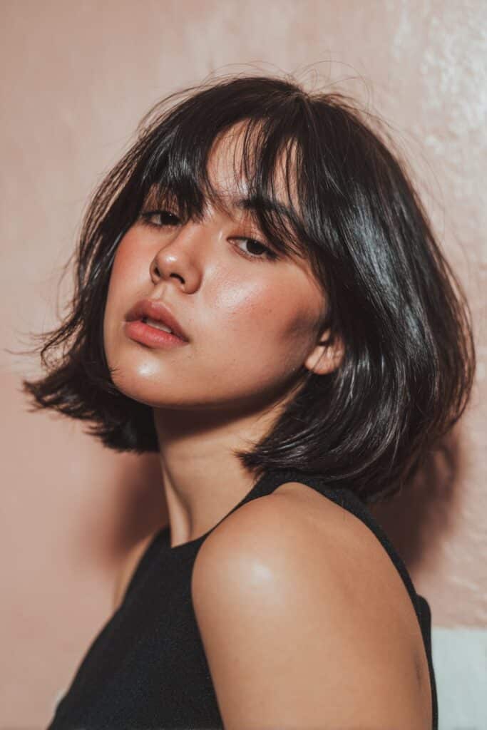 A stunning young woman in her 20s with natural skin tone, showcasing a bob hairstyle for going out featuring a bob with curtain bangs, featuring a bob with curtain-style bangs, captured in a close-up shot focusing on the hair, photographed against a seamless deep teal backdrop with a smooth texture for optimal color contrast. She has subtle makeup, a chic evening outfit, realistic hair texture, warm skin tone, and a confident expression, captured with soft natural lighting and an Instagram-style beauty vibe.