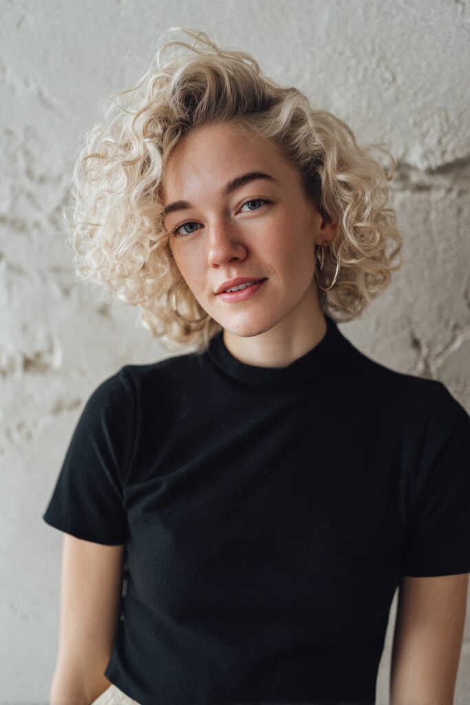 Woman with short half-up Bardot bouffant, waves, platinum blonde hair, plaster backdrop.