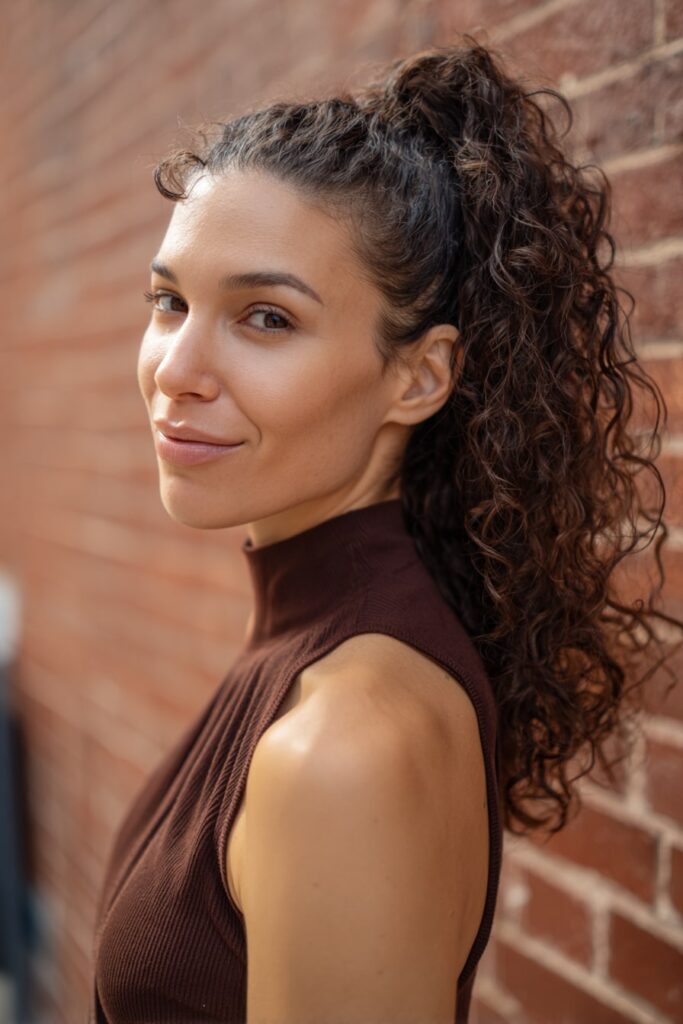 Woman with half up half down curly hair, bubble ponytail, curly ends, brick backdrop.