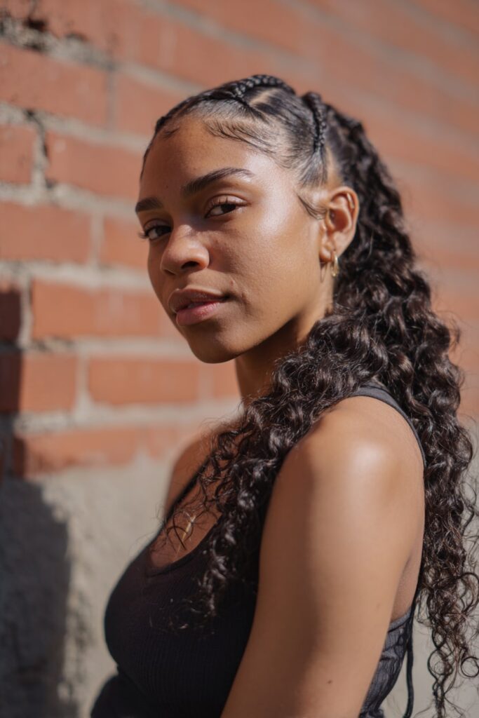 Black girl with half-up butterfly braids, curly braiding hair, dark brown, brick backdrop.
