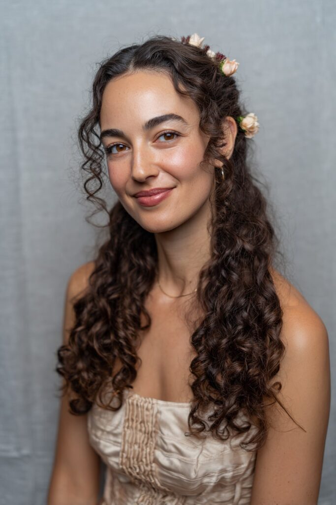 Woman with half up half down curly hair, cascading curls, floral pins, gray backdrop.