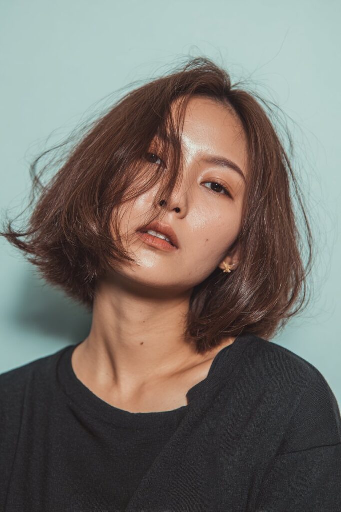 A stunning young woman in her 20s with natural skin tone, showcasing a bob hairstyle with side bangs featuring a choppy bob with long side bangs, featuring a choppy bob with long, side-swept bangs, captured in a close-up shot focusing on the hair, photographed against a seamless light teal backdrop with a glossy sheen. She has subtle makeup, a chic outfit, realistic hair texture, warm skin tone, and a confident expression, captured with soft natural lighting and an Instagram-style beauty vibe.