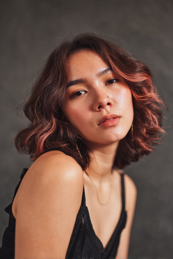 A stunning young woman in her 20s with natural skin tone, showcasing a bob hairstyle for going out featuring a colored bob, featuring a bob with vibrant colored highlights, captured in a close-up shot focusing on the hair, photographed against a seamless deep charcoal backdrop with a glossy texture for optimal color contrast. She has subtle makeup, a chic evening outfit, realistic hair texture, warm skin tone, and a confident expression, captured with soft natural lighting and an Instagram-style beauty vibe.