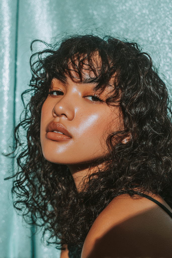 A stunning young woman in her 20s with natural skin tone, showcasing a homecoming hairstyle for curly hair with a curly bangs moment, featuring curly bangs framing the face with loose curls, captured in a close-up shot focusing on the hair, photographed against a seamless light teal backdrop with a glossy sheen. She has subtle makeup, a chic homecoming dress, realistic hair texture, warm skin tone, and a confident expression, captured with soft natural lighting and an Instagram-style beauty vibe.