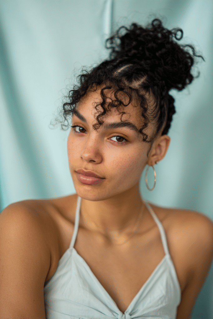 A stunning young woman in her 20s with natural skin tone, showcasing a homecoming hairstyle for curly hair with a curly crown braid, featuring a crown braid made with curly hair, captured in a close-up shot focusing on the hair, photographed against a seamless soft mint backdrop with a silky texture. She has subtle makeup, a chic homecoming dress, realistic hair texture, warm skin tone, and a confident expression, captured with soft natural lighting and an Instagram-style beauty vibe.