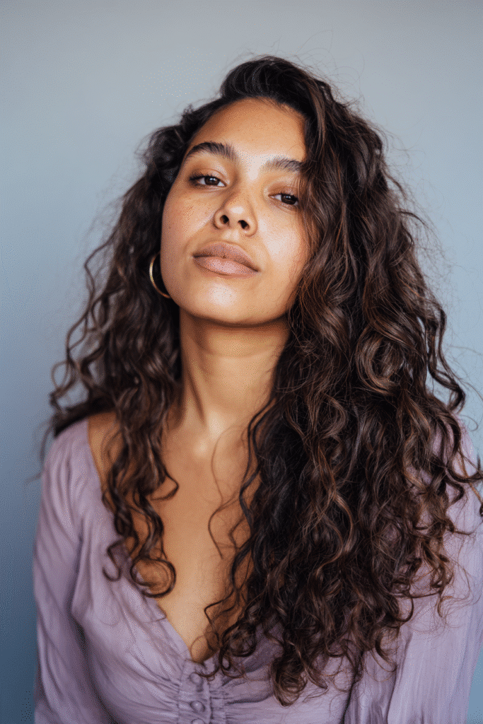 A stunning young woman in her 20s with natural skin tone, showcasing a homecoming hairstyle for curly hair with defined & down curls, featuring defined, flowing curls left down, captured in a close-up shot focusing on the hair, photographed against a seamless soft lavender backdrop with a matte finish. She has subtle makeup, a chic homecoming dress, realistic hair texture, warm skin tone, and a confident expression, captured with soft natural lighting and an Instagram-style beauty vibe.